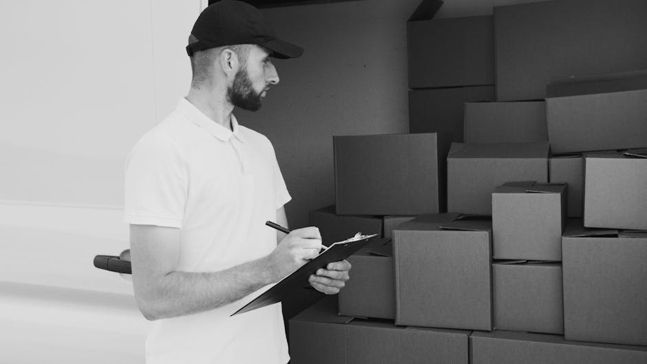 A man with a beard and wearing a black cap and white polo shirt stands indoors beside a stack of uniform cardboard moving boxes. He is holding a clipboard and a pen, appearing to take notes or check off items during a home relocation process. The boxes are neatly arranged against a wall in a room with plain walls, and some are stacked on top of each other at varying heights. The lighting is even and natural, indicating a professional environment focused on packing and moving logistics, with the man representing a mover or coordinator from Barnet Removals assessing the load in preparation for furniture transport or a house move.
