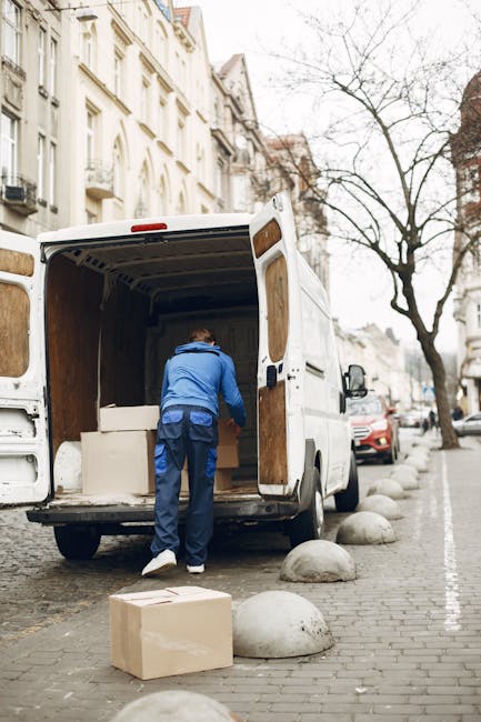 A mover wearing a blue jacket and dark trousers is inside the open cargo area of a white moving van, arranging or loading large cardboard boxes and other packing materials. The van is parked on a city street with cobblestone paving, next to a row of rounded concrete road barriers. Nearby, a cardboard box is placed on the pavement, and several parked cars are visible along the street. In the background, there are multi-storey residential buildings with ornate facades and a leafless tree, indicating a potential late autumn or winter setting. The scene captures the process of home relocation, with visible furniture and packing supplies inside the van, as part of a professional removals service offered by Barnet Removals. Natural daylight illuminates the area, emphasizing the urban environment and the logistics involved in furniture transport and moving logistics.