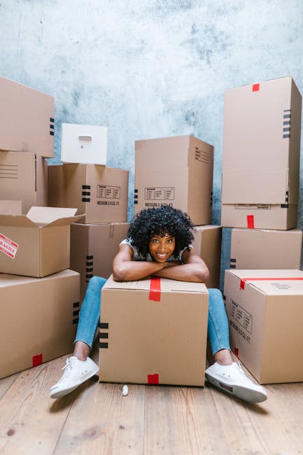 A young woman with curly black hair, wearing a white t-shirt, blue jeans, and white sneakers, is sitting on the floor in a room filled with numerous cardboard moving boxes of various sizes, some sealed with red or black packaging tape. She is smiling and resting her chin on her forearms, which are placed on a large cardboard box in front of her. The boxes are arranged around her, some placed directly on the wooden floor, which has a light natural finish. The background features a textured blue and white wall, creating a neutral setting suitable for showcasing home relocation or packing services, as operated by Barnet Removals. The image captures the process of packing and preparing for a house move, emphasizing the handling and organization of belongings before transportation, aligned with removal service themes.