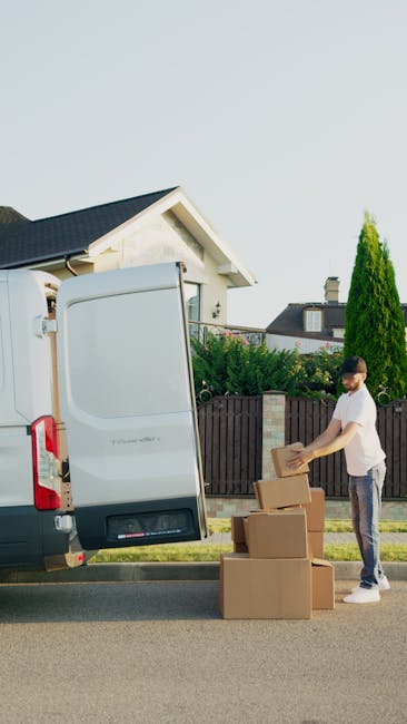 A young man with short dark hair and a beard, wearing a light grey t-shirt and dark trousers, is seen standing on the pavement beside a white removal van with its rear door open. He is loading or unloading medium-sized cardboard boxes, some of which are on the ground in front of him, while others are placed inside the van. The boxes are sealed with packing tape and made of brown corrugated cardboard. The removal vehicle is parked outside a residential property, with a black wooden fence, green bushes, and a tall, narrow conifer tree visible in the background. The house has a white exterior with a gabled roof covered in dark tiles, and the scene is illuminated by natural daylight, suggesting a clear weather condition. Barnet Removals, a professional moving service, appears to be carrying out a home relocation or furniture transport process, involving packing and loading materials used to facilitate efficient house removals.