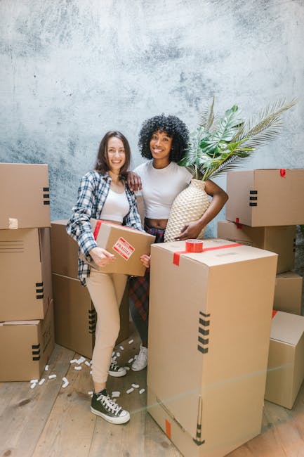 A young woman with curly black hair, wearing a white t-shirt, blue jeans, and white sneakers, is sitting on the floor in a room filled with numerous cardboard moving boxes of various sizes, some sealed with red or black packaging tape. She is smiling and resting her chin on her forearms, which are placed on a large cardboard box in front of her. The boxes are arranged around her, some placed directly on the wooden floor, which has a light natural finish. The background features a textured blue and white wall, creating a neutral setting suitable for showcasing home relocation or packing services, as operated by Barnet Removals. The image captures the process of packing and preparing for a house move, emphasizing the handling and organization of belongings before transportation, aligned with removal service themes.