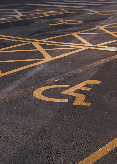 Close-up view of a black asphalt parking lot surface with white painted markings indicating accessible parking spaces. The largest symbol is a wheelchair icon within a rectangular border, positioned n